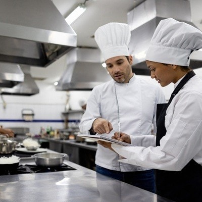 Chef talking to a student in a cooking class while she writes it down a recipe on a notebook